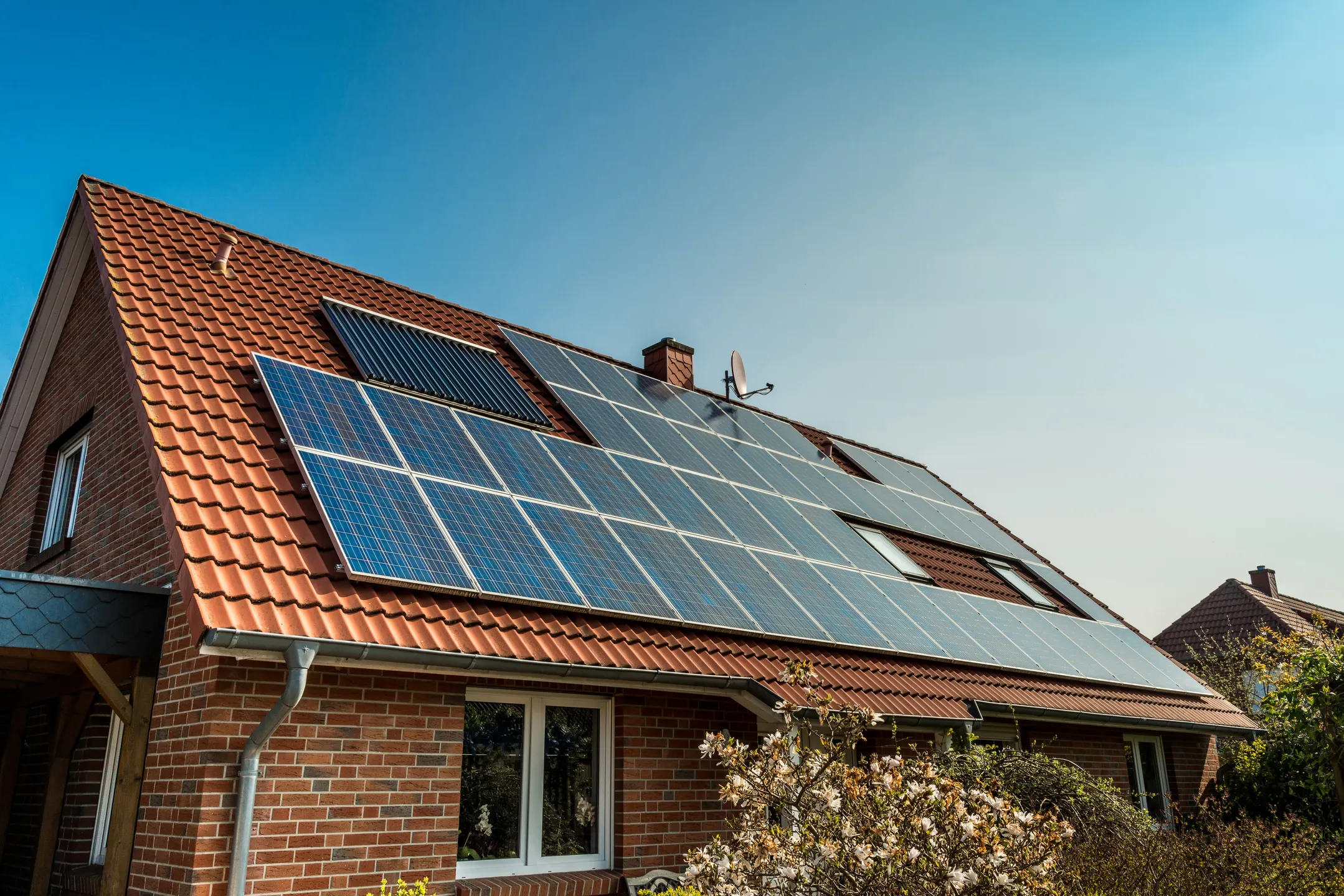 A red brick house with a sloped, tiled roof is equipped with several solar panels. The panels, which undergo regular solar panel maintenance, cover most of the roof's surface, capturing sunlight. A small satellite dish is also visible on the roof. Trees in the foreground are partially in bloom.