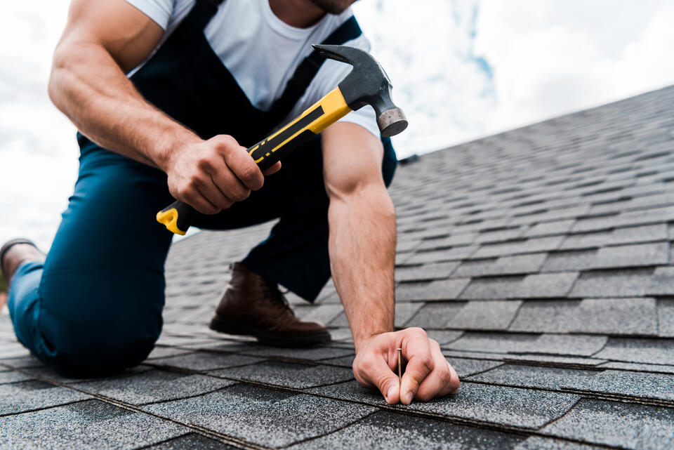 roofing tools-close up of man in blue overalls using hammer to install roof shingles on a house